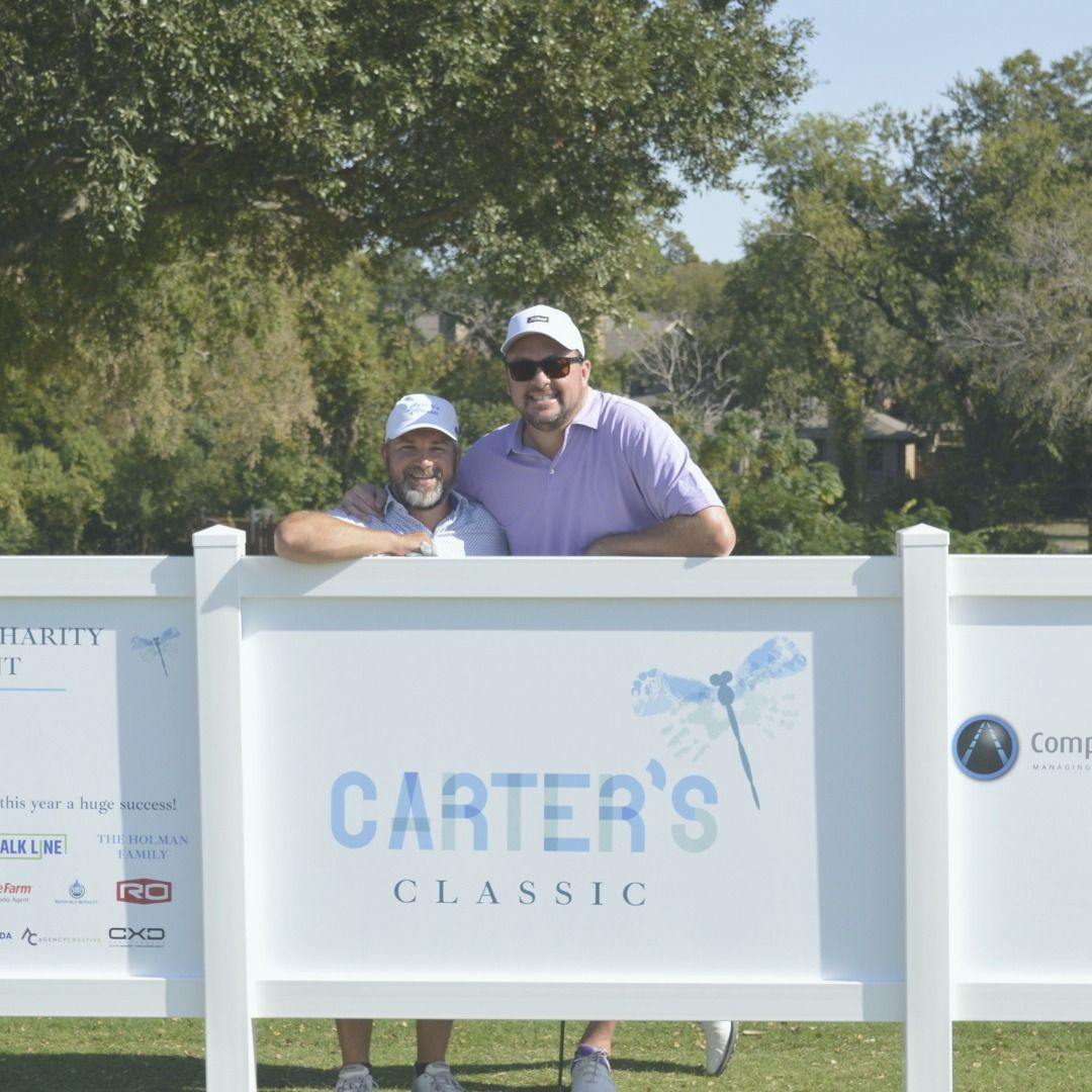 Two people smiling behind a white Carter’s Classic golf sign outdoors on a sunny day