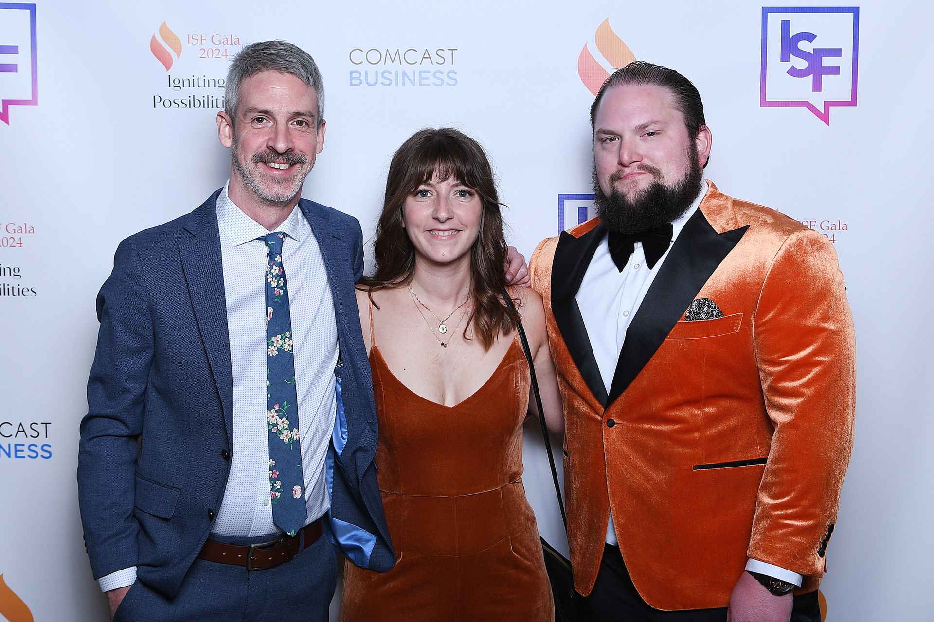 Three people posing at a step-and-repeat event, with one in a blue suit and two in orange outfits.