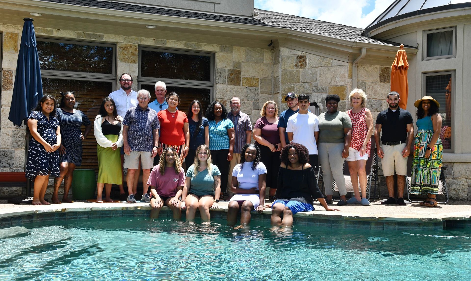 Group of people posing by a backyard swimming pool on a sunny day, with a stone house in the background