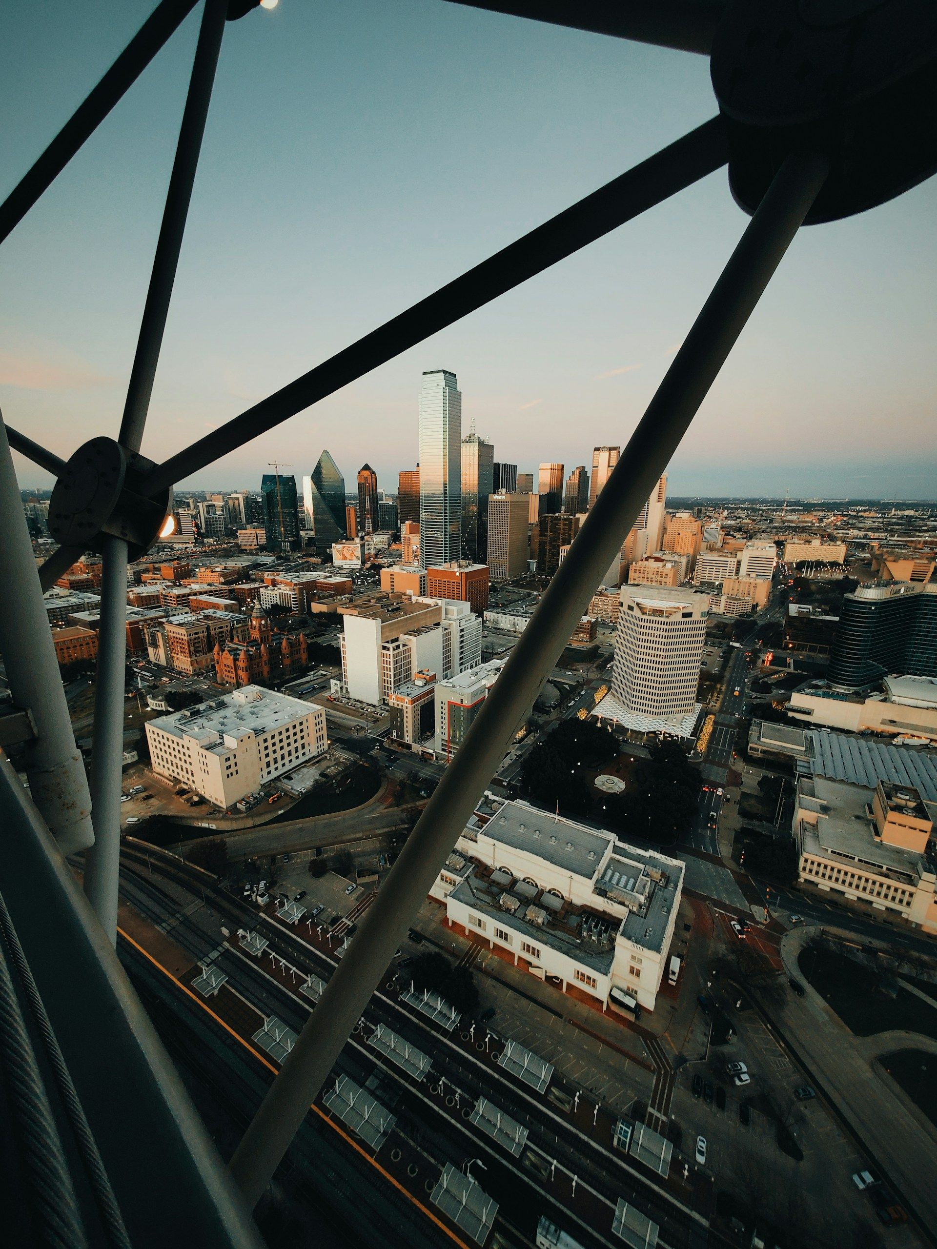 City skyline at sunset viewed through dark metal beams, with roads and buildings below.