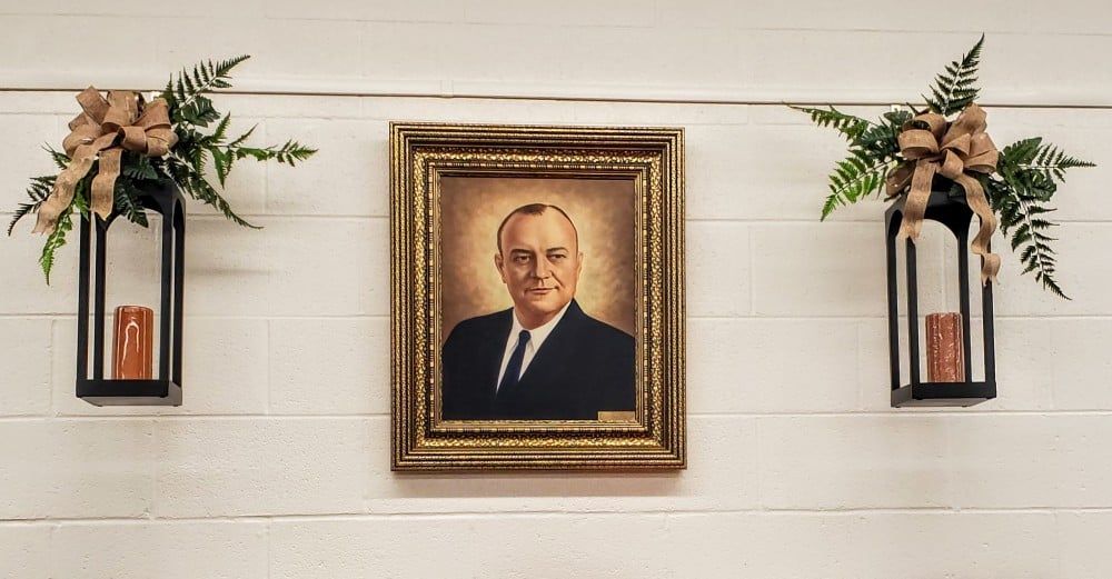 Portrait in ornate frame, flanked by lanterns with greenery, on a white plank wall.