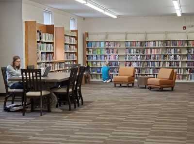 Library interior with bookshelves, tables, chairs, and two people reading. Brown and tan carpet.