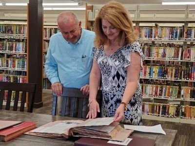 Man and woman examining a book at a library table, bookshelves in the background.