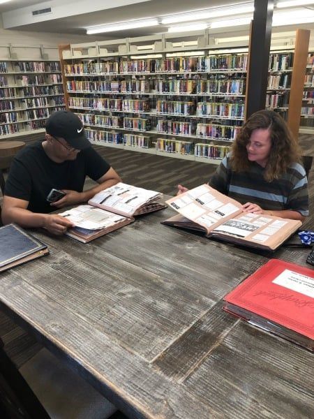 Two people at a table in a library looking at open books. Bookshelves in the background.