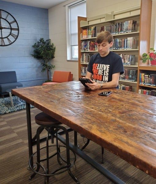 Person reading an e-reader at a large wooden table in a library. Bookshelves in the background.