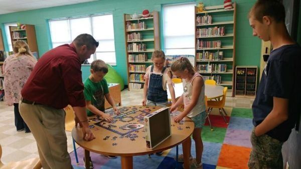 People at a library table working on a jigsaw puzzle, shelves of books in the background.