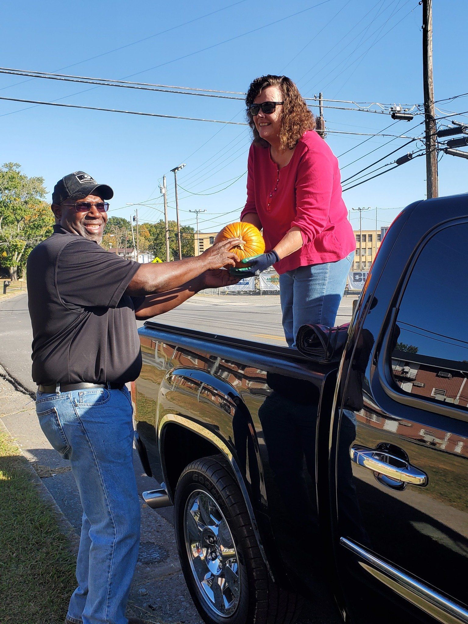 Man hands a pumpkin to a woman standing in a black pickup truck bed on a sunny day.