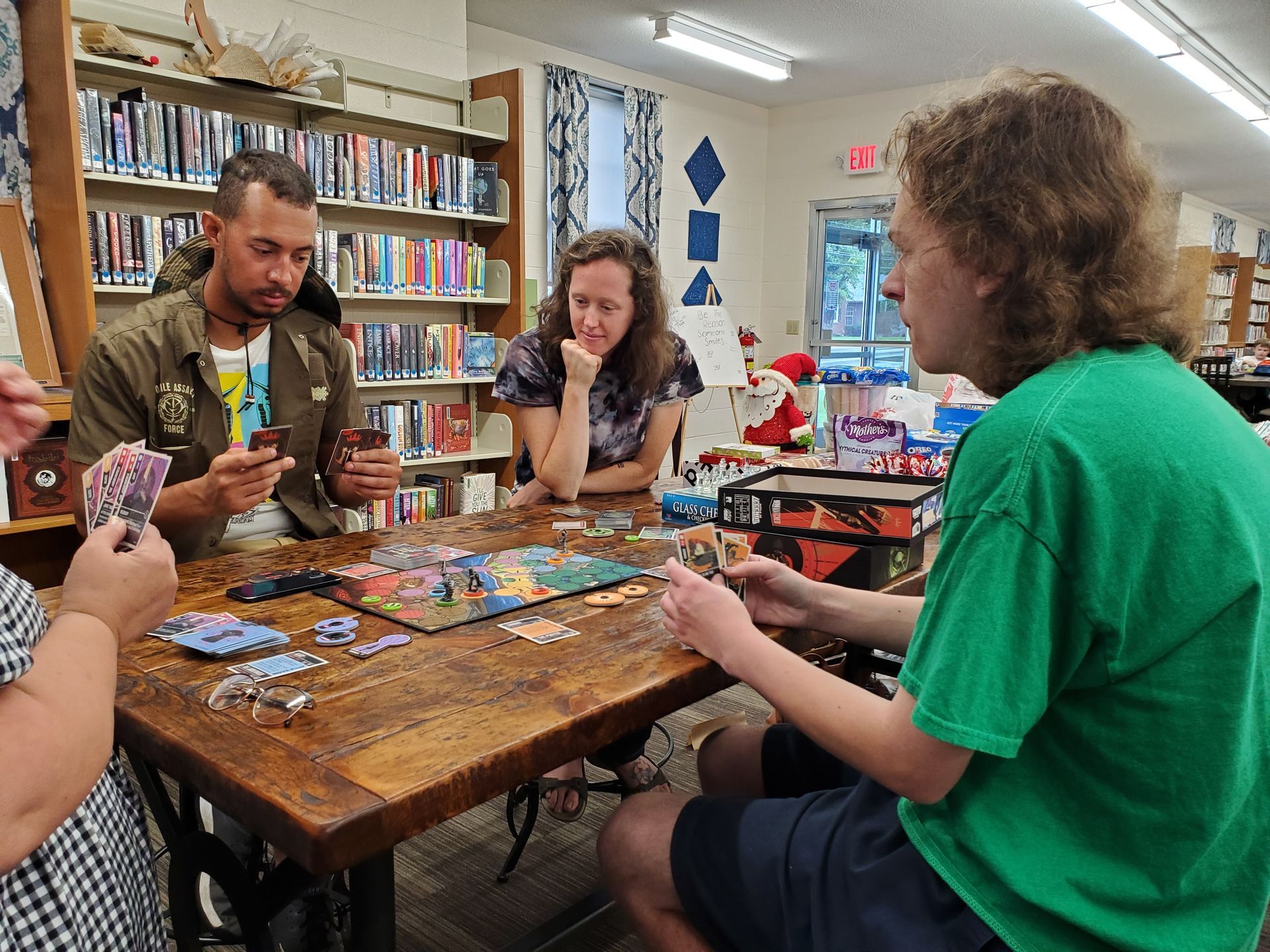 People playing a board game at a table in a library, surrounded by bookshelves.