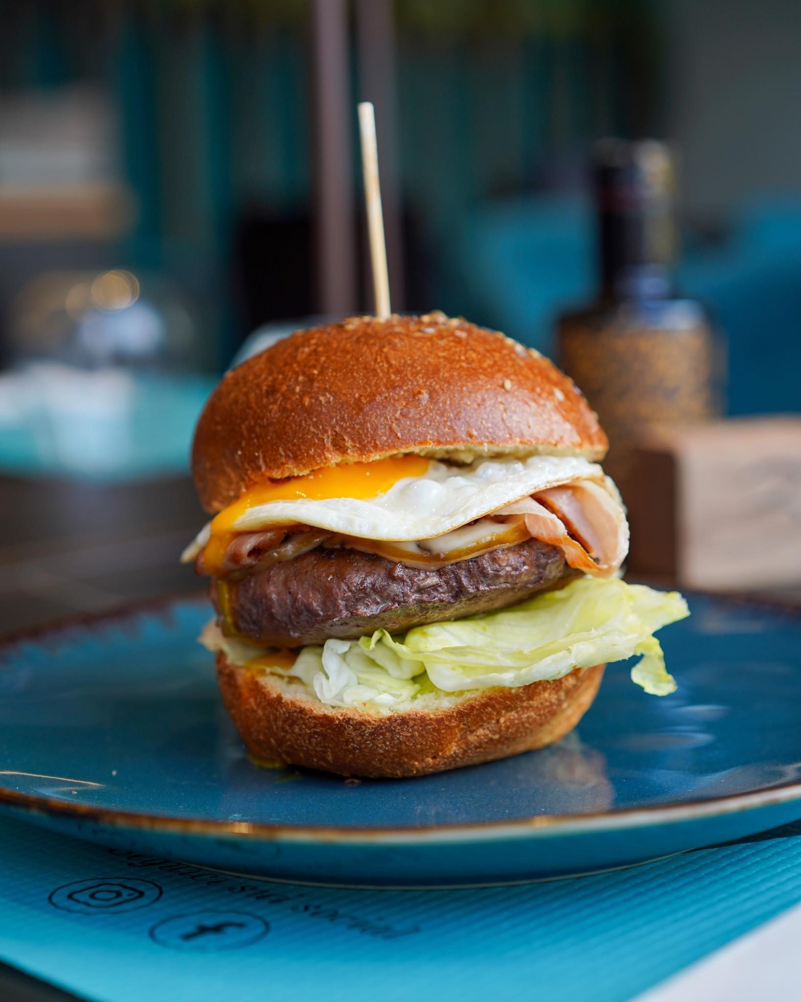 Hamburger servito su un piatto blu, guarnito con uovo fritto e pancetta, il tutto tenuto insieme da uno spiedino.