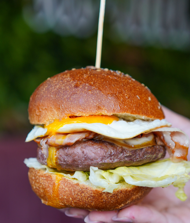 Hamburger con uovo e pancetta su un panino brioche, tenuto in mano e infilzato con uno spiedino.