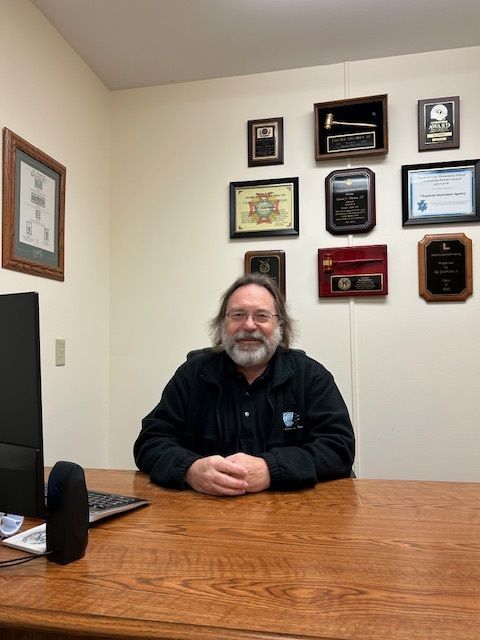 A man is sitting at a desk in a room with plaques on the wall.