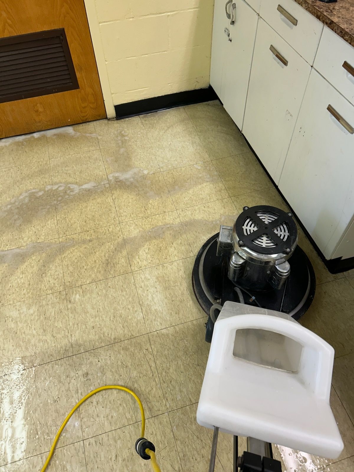 A floor buffer cleans a beige tiled floor in a room with white cabinets and a wooden door.