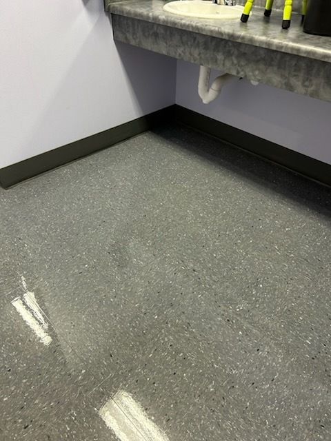 A corner of a room with speckled grey vinyl flooring, dark baseboards, and a wall-mounted sink with exposed plumbing.