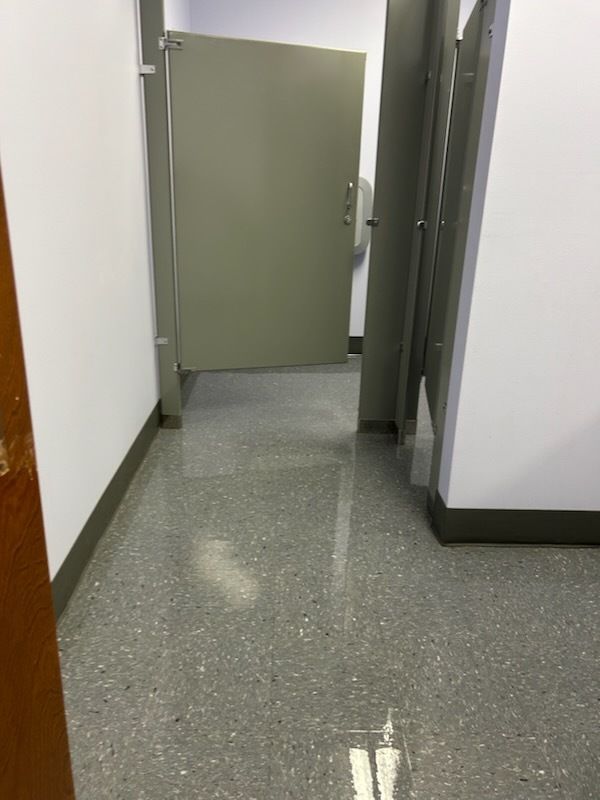 A commercial restroom with light gray stalls and a shiny, wet floor, indicating a potential spill or cleaning in progress.
