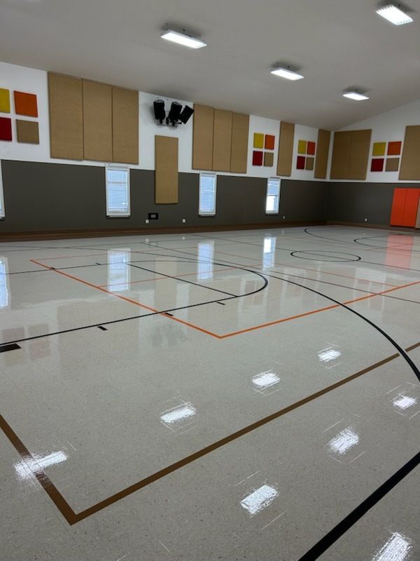 A brightly lit indoor gymnasium with a glossy light-colored floor featuring orange and black game court lines.