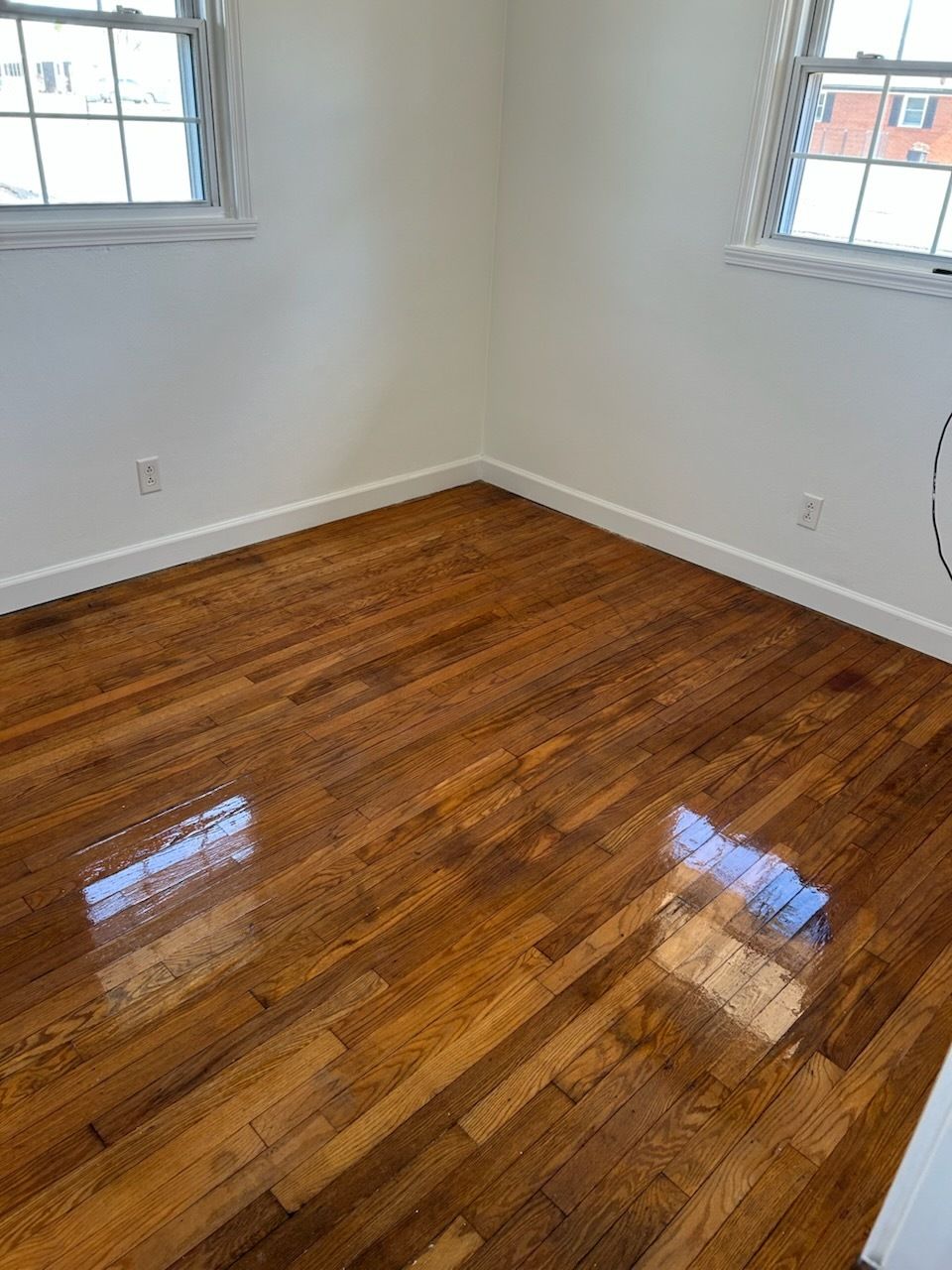Empty room with polished hardwood floors, white walls, and two windows reflecting daylight.