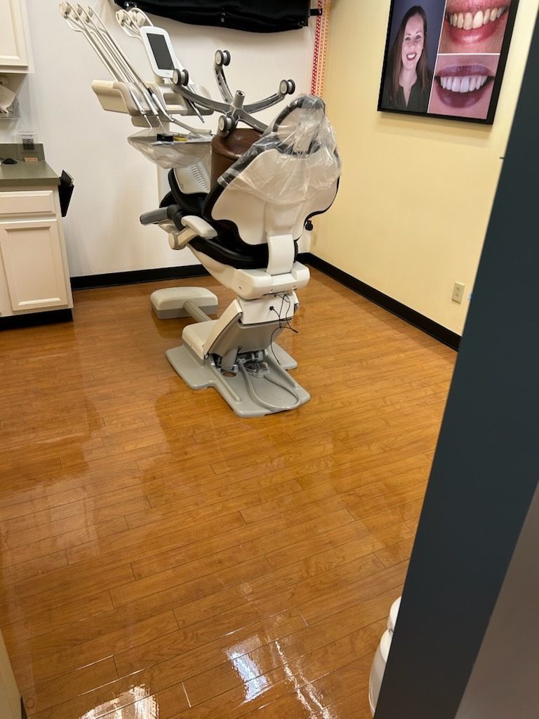 A dental exam chair with equipment in a clinical office with light-colored walls and wood-patterned flooring.