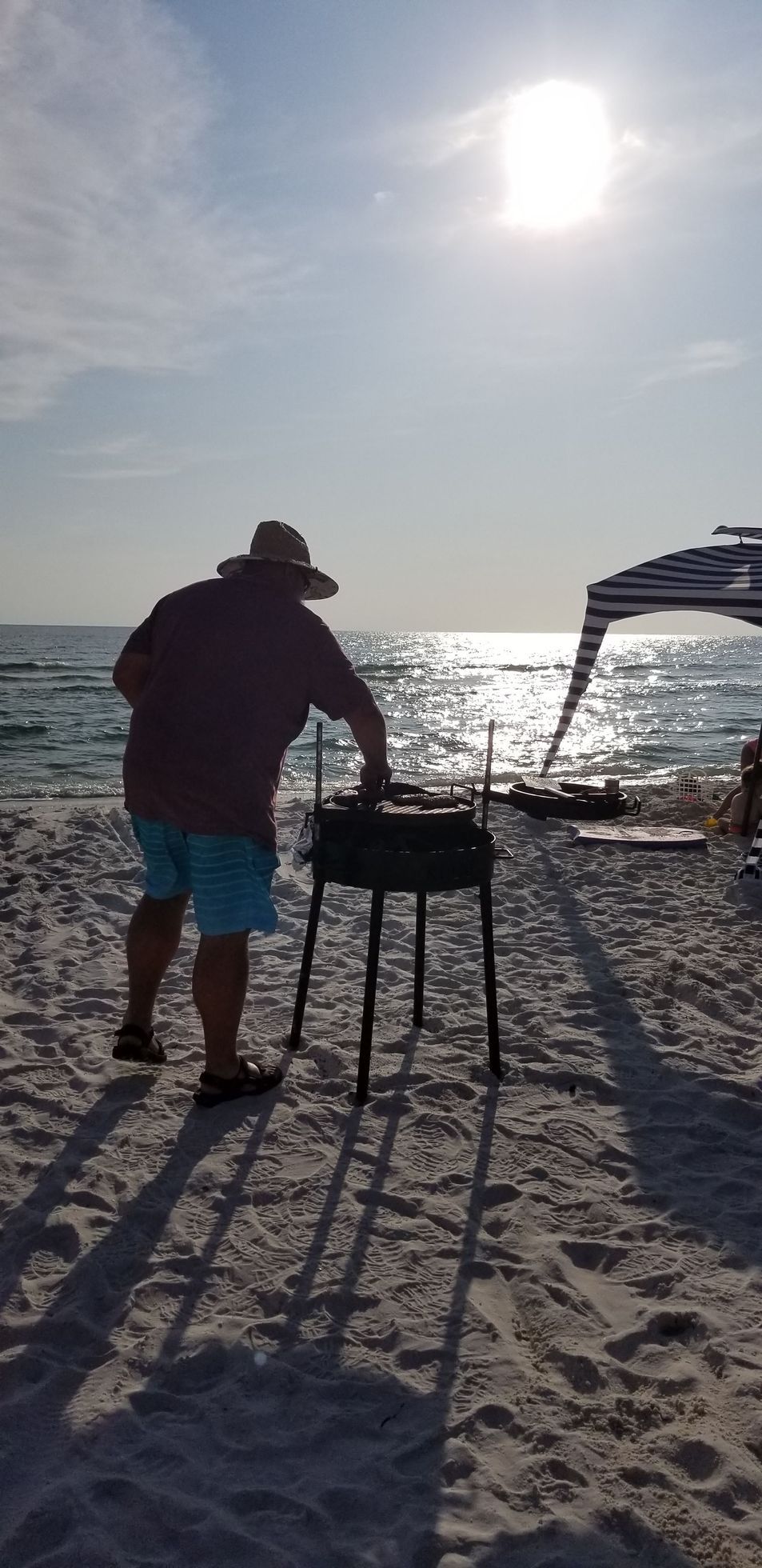Man grilling on a beach at sunset, with the sea and a sun umbrella in the background.
