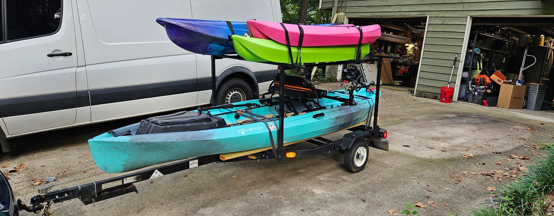 A trailer carrying three kayaks in various colors: purple, pink, green, and turquoise. Beside a white van.
