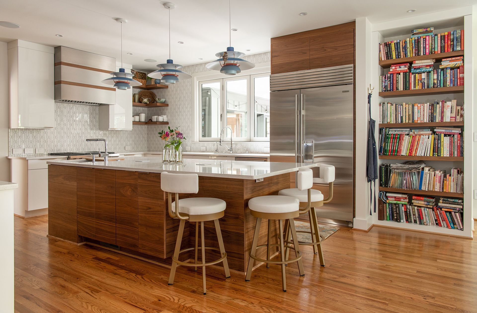 Modern kitchen with wooden island and stools, stainless steel fridge, built-in bookshelf.