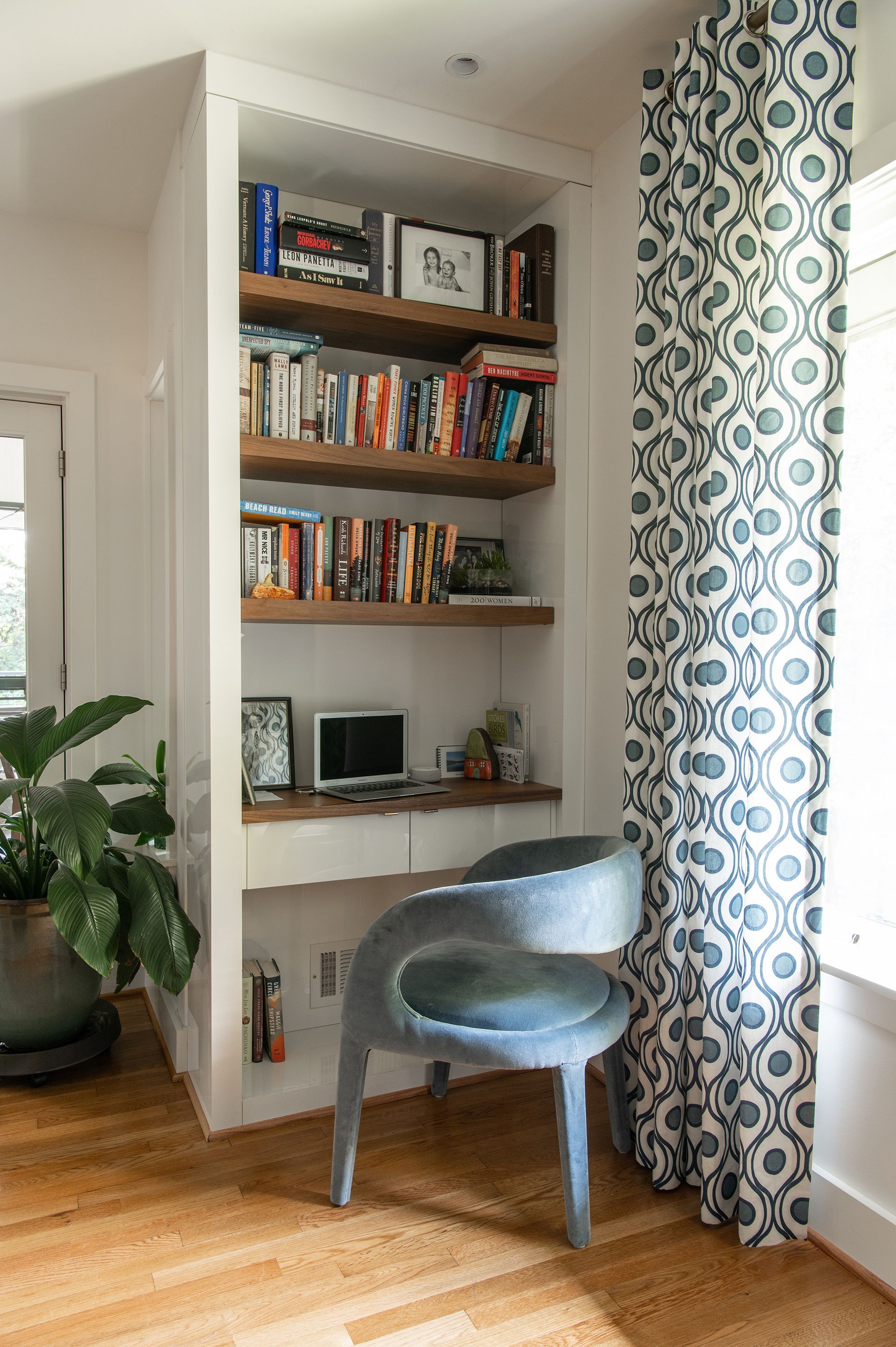 Built-in bookshelf with desk and blue chair. White walls, wooden shelves, and patterned curtains.