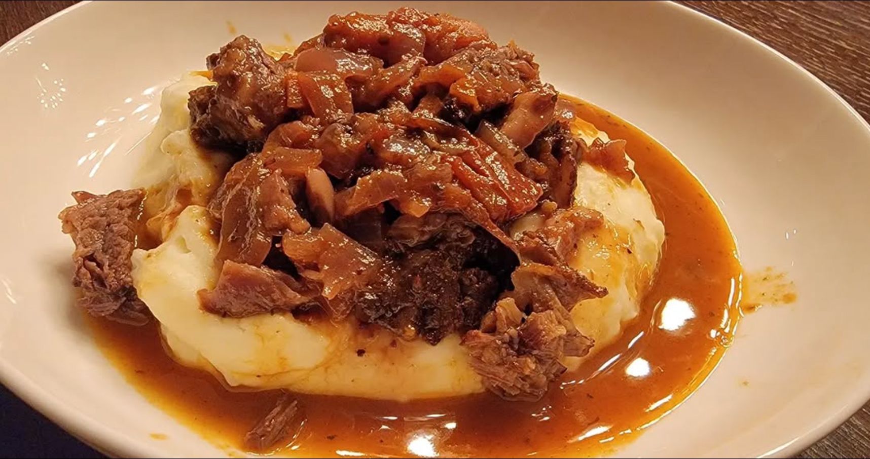 Mashed potatoes topped with braised meat and gravy in a white bowl.