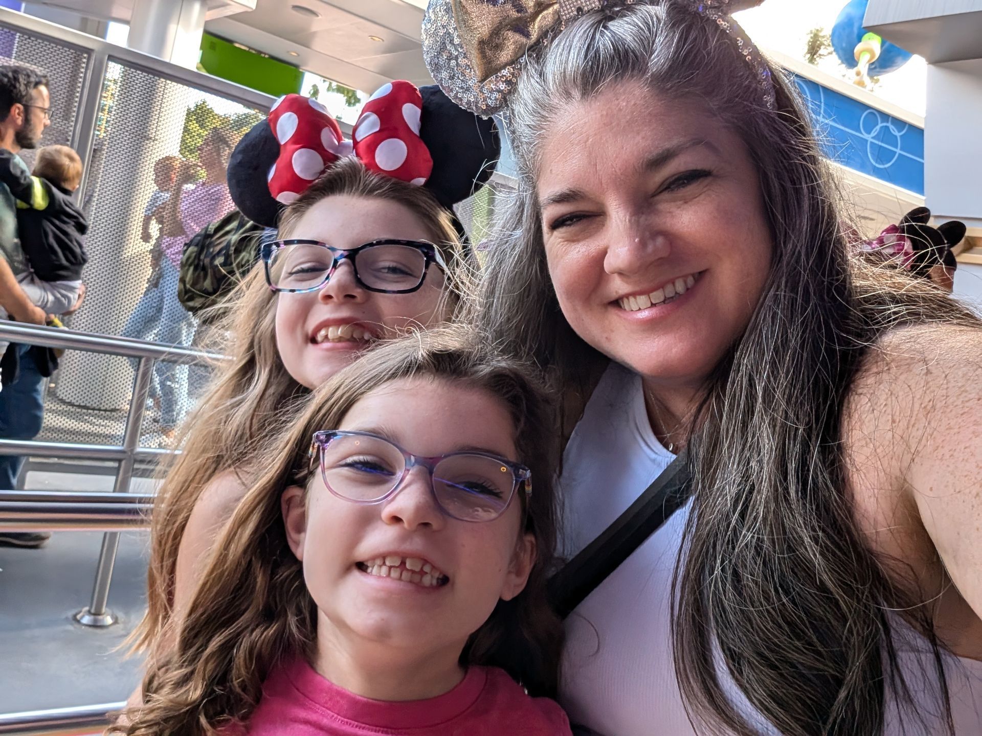Woman and two children smile, wearing Minnie Mouse ears and glasses, in a theme park.