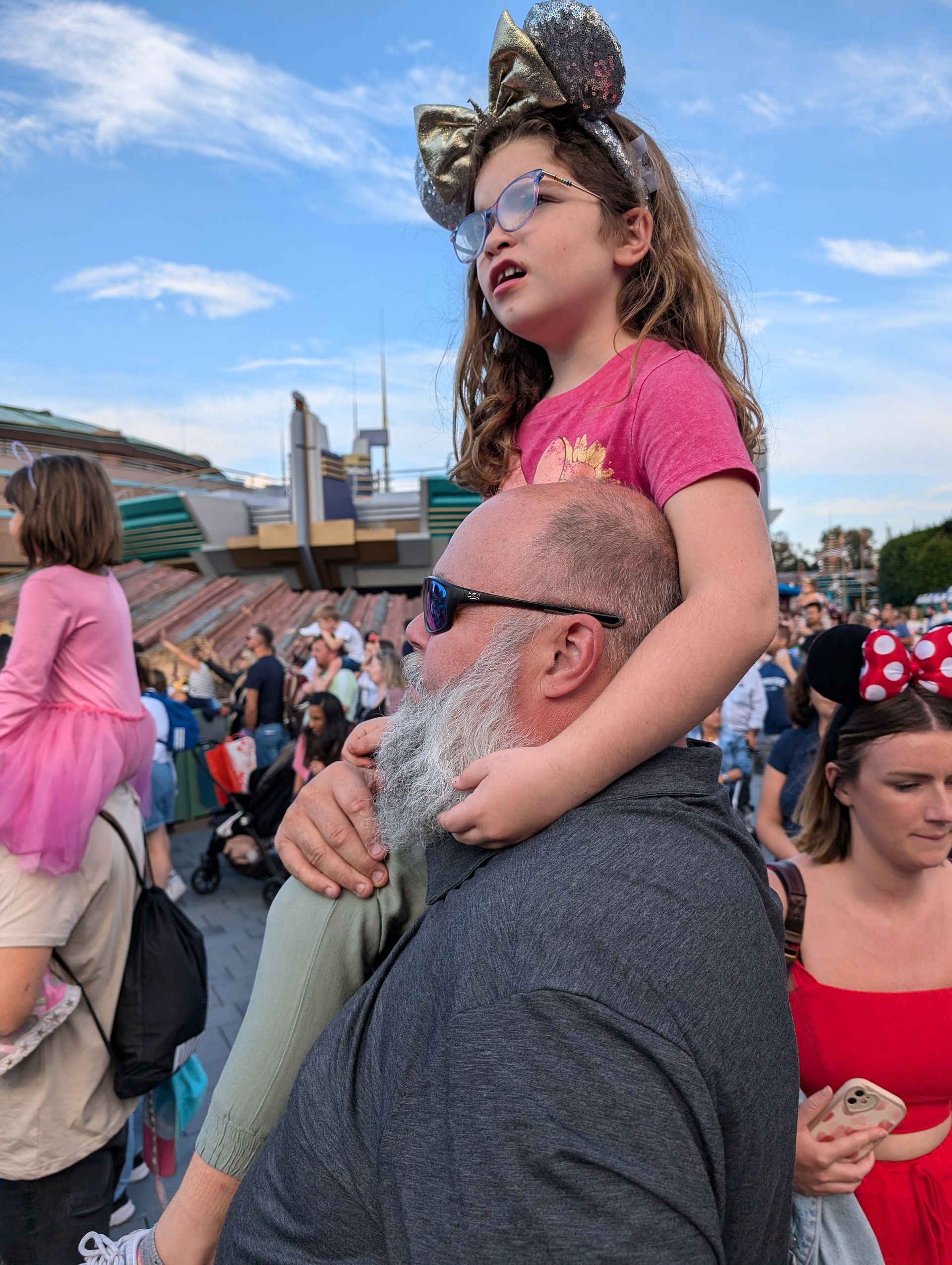 A child wearing Mickey Mouse ears sits on a person's shoulders in a crowd, outdoors, at a theme park.