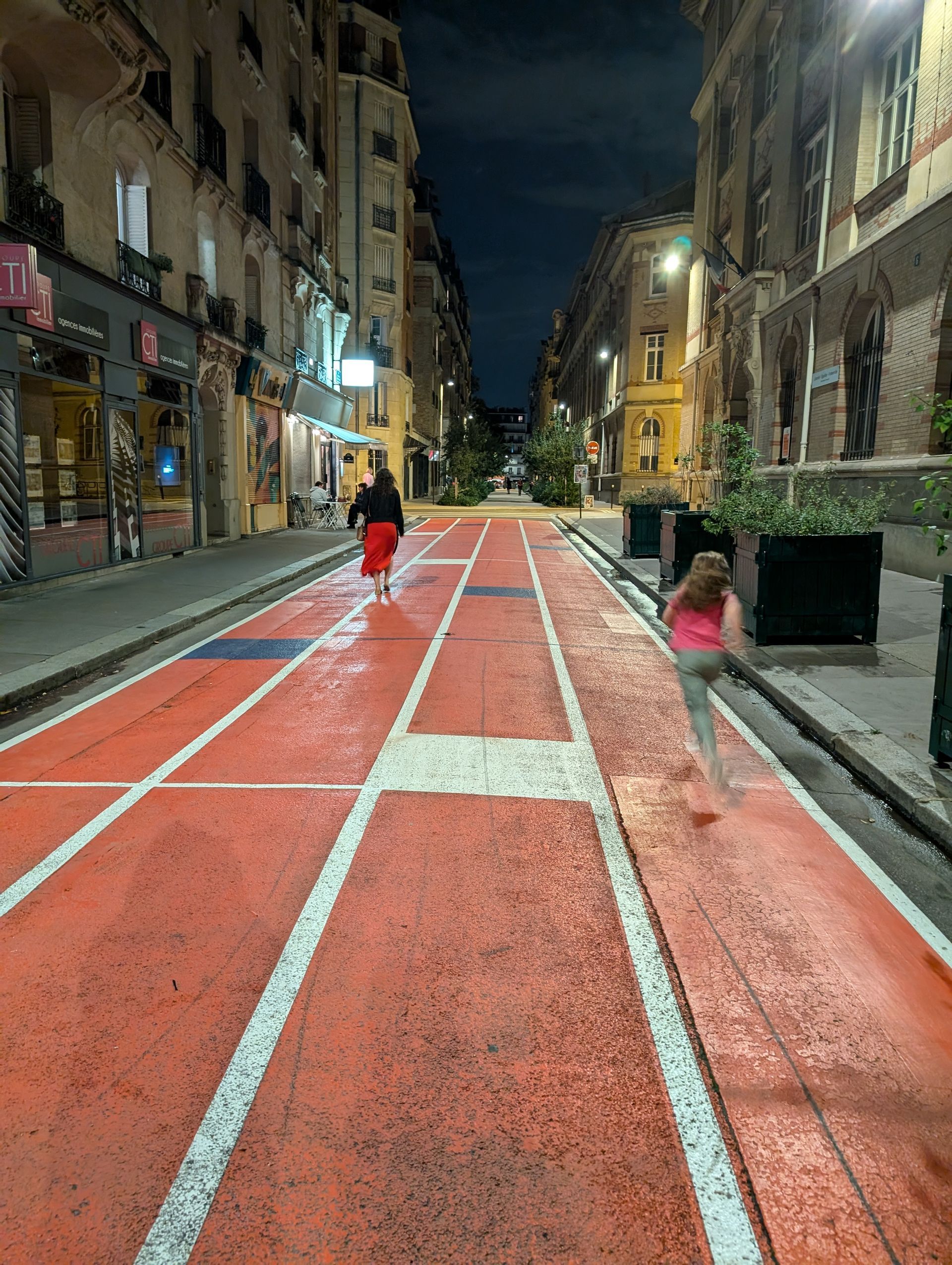 People running on a red painted street designed as a track. Buildings line both sides. Night scene.