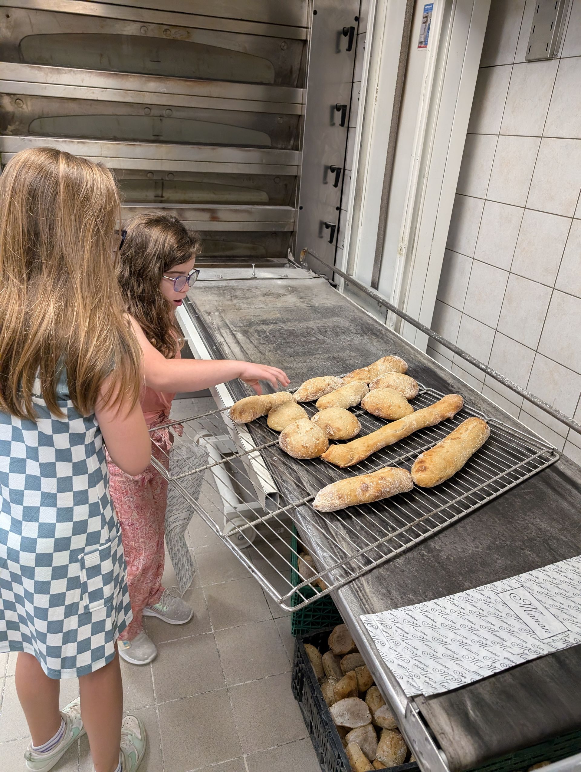 Two children examining freshly baked bread on a conveyor belt in a bakery.