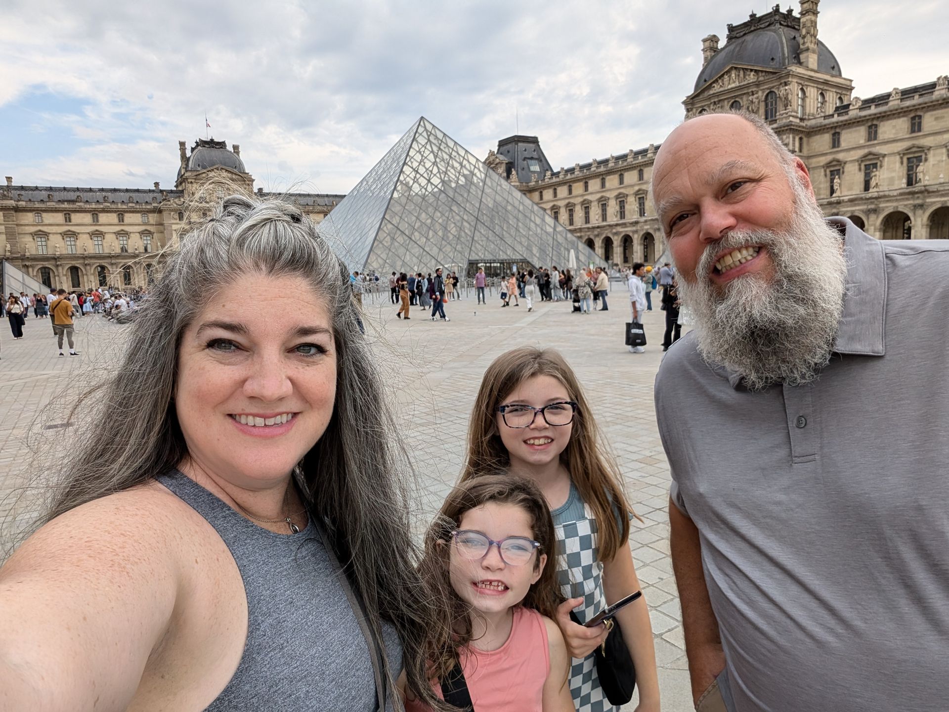 Family selfie in front of the Louvre Museum in Paris, with glass pyramid and people in background.