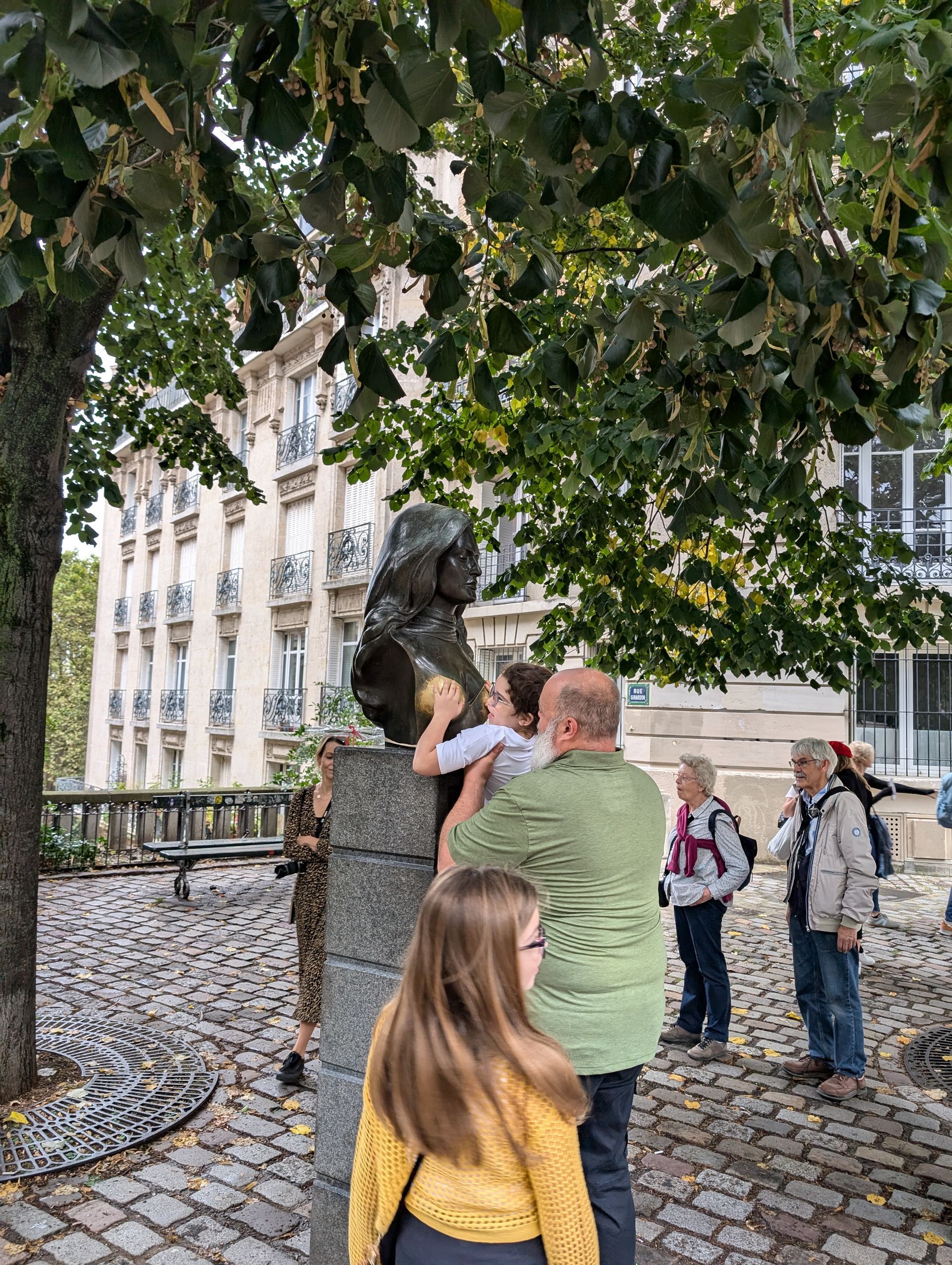 People near a sculpture under a tree. One person touches the sculpture's head. Others watch nearby on a cobblestone path.