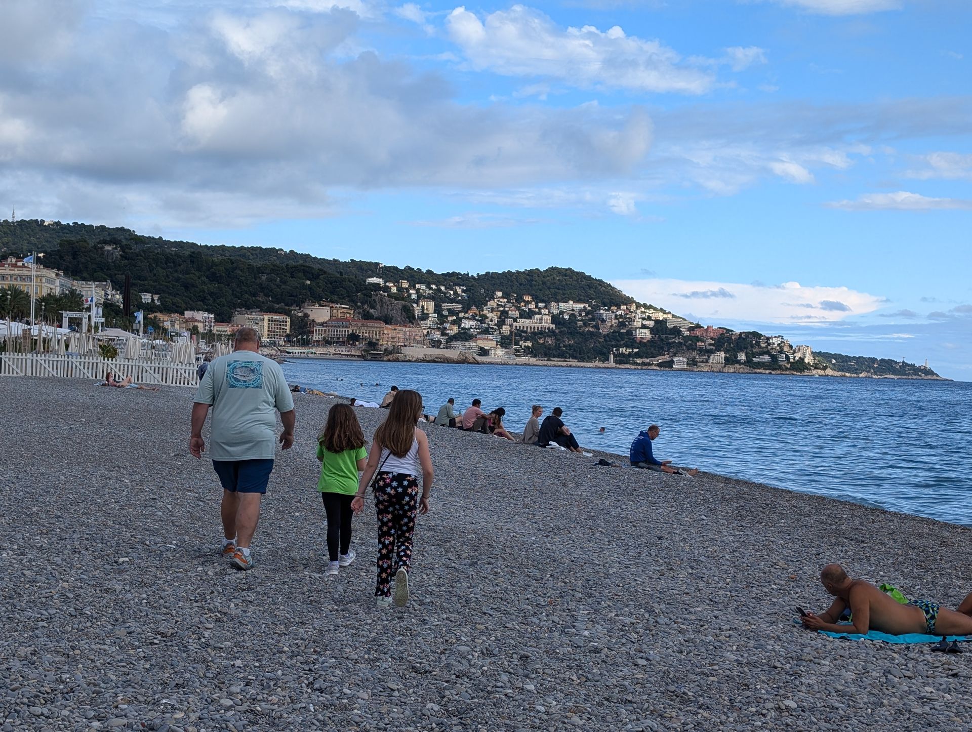 Family walks along a pebbled beach in Nice, France, toward the sea under a cloudy sky.