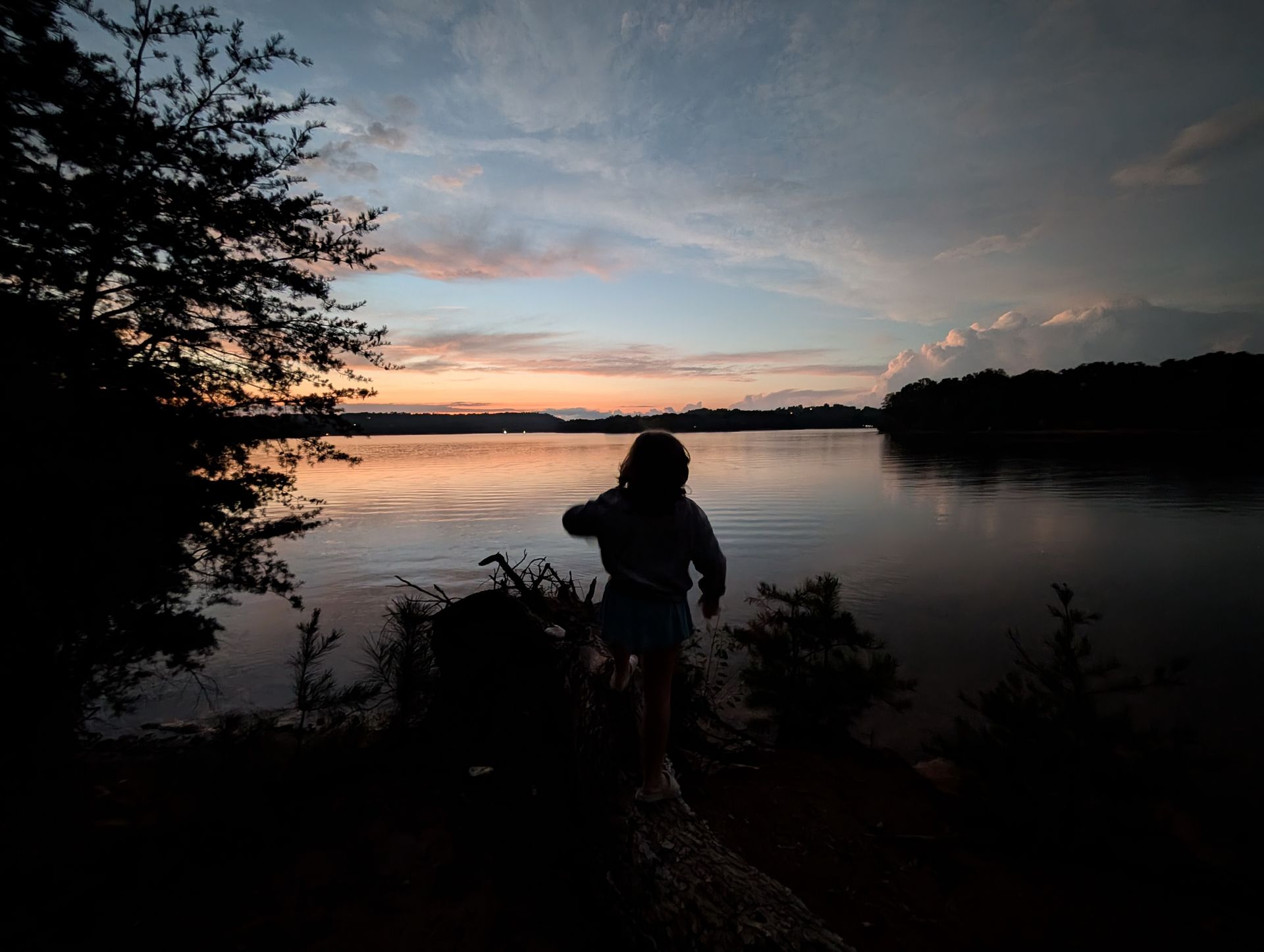Silhouette of person throwing a stone into a lake at sunset. Dark trees frame the water and colorful sky.
