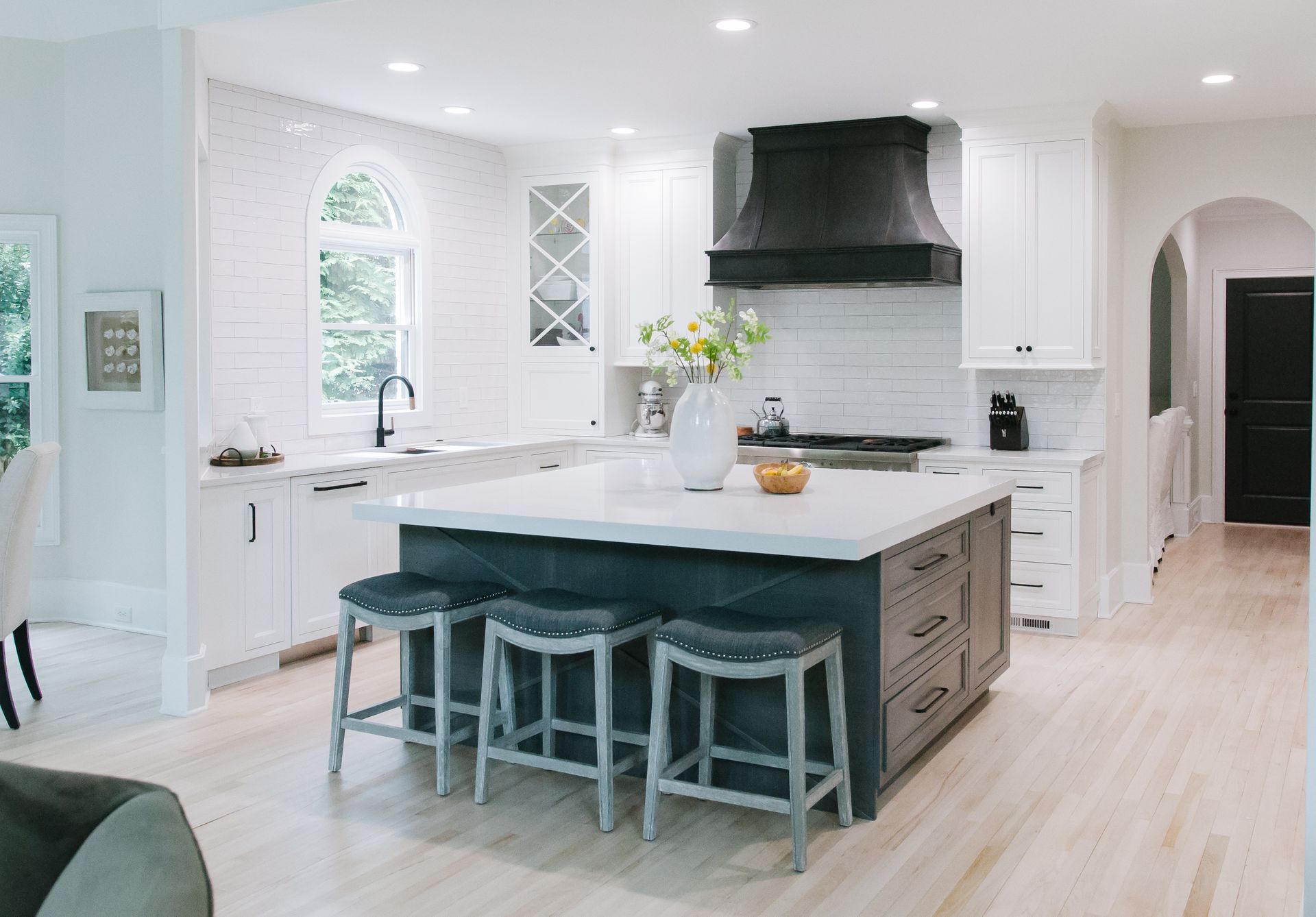 Kitchen with white cabinets, dark island, black hood, and gray bar stools.