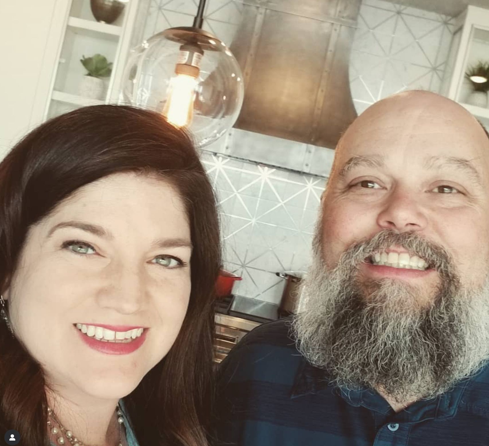 Smiling couple taking a selfie in a modern kitchen. She has dark hair, he has a beard.