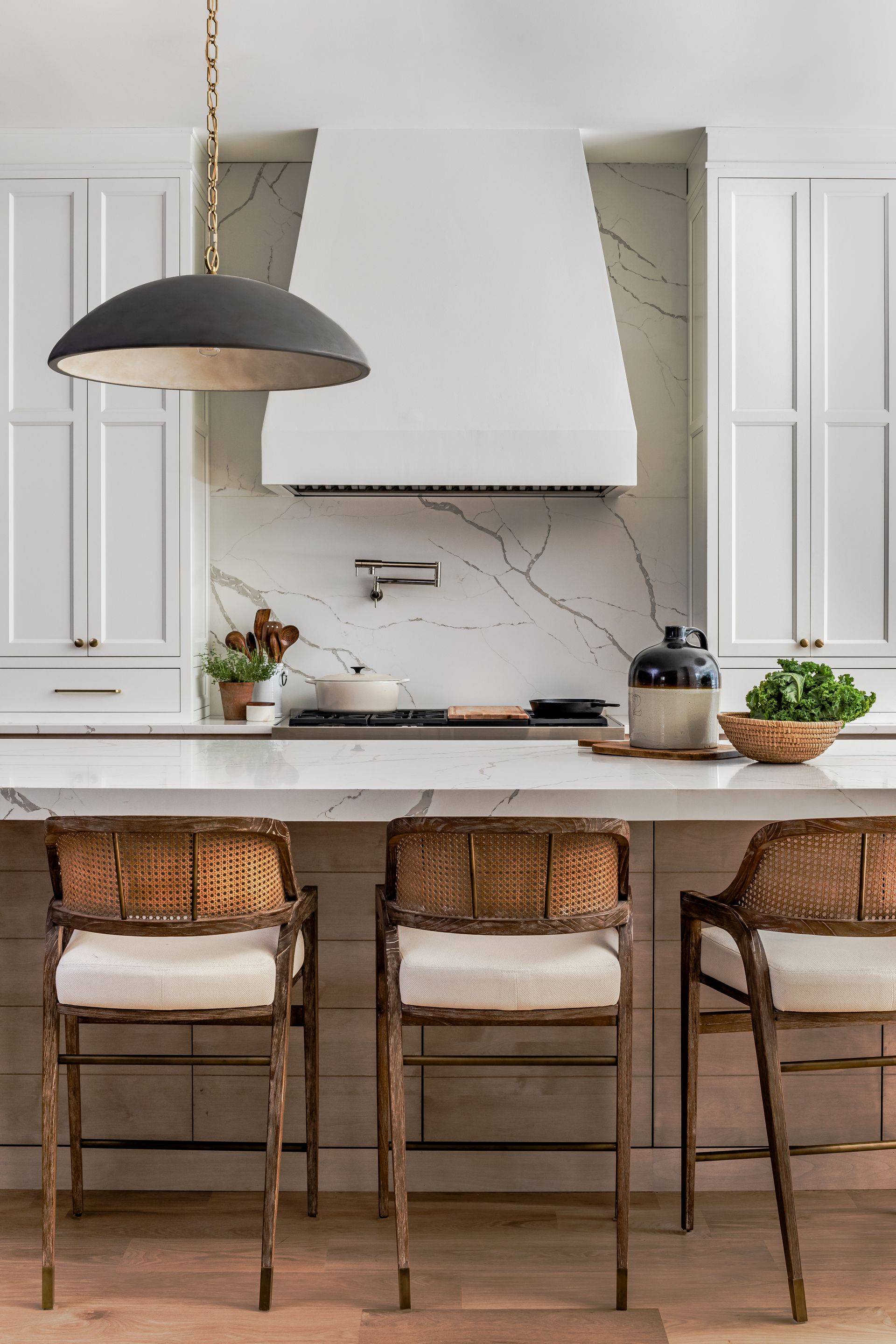 Kitchen with white cabinets, marble backsplash, dark gray pendant light, and wicker bar stools.