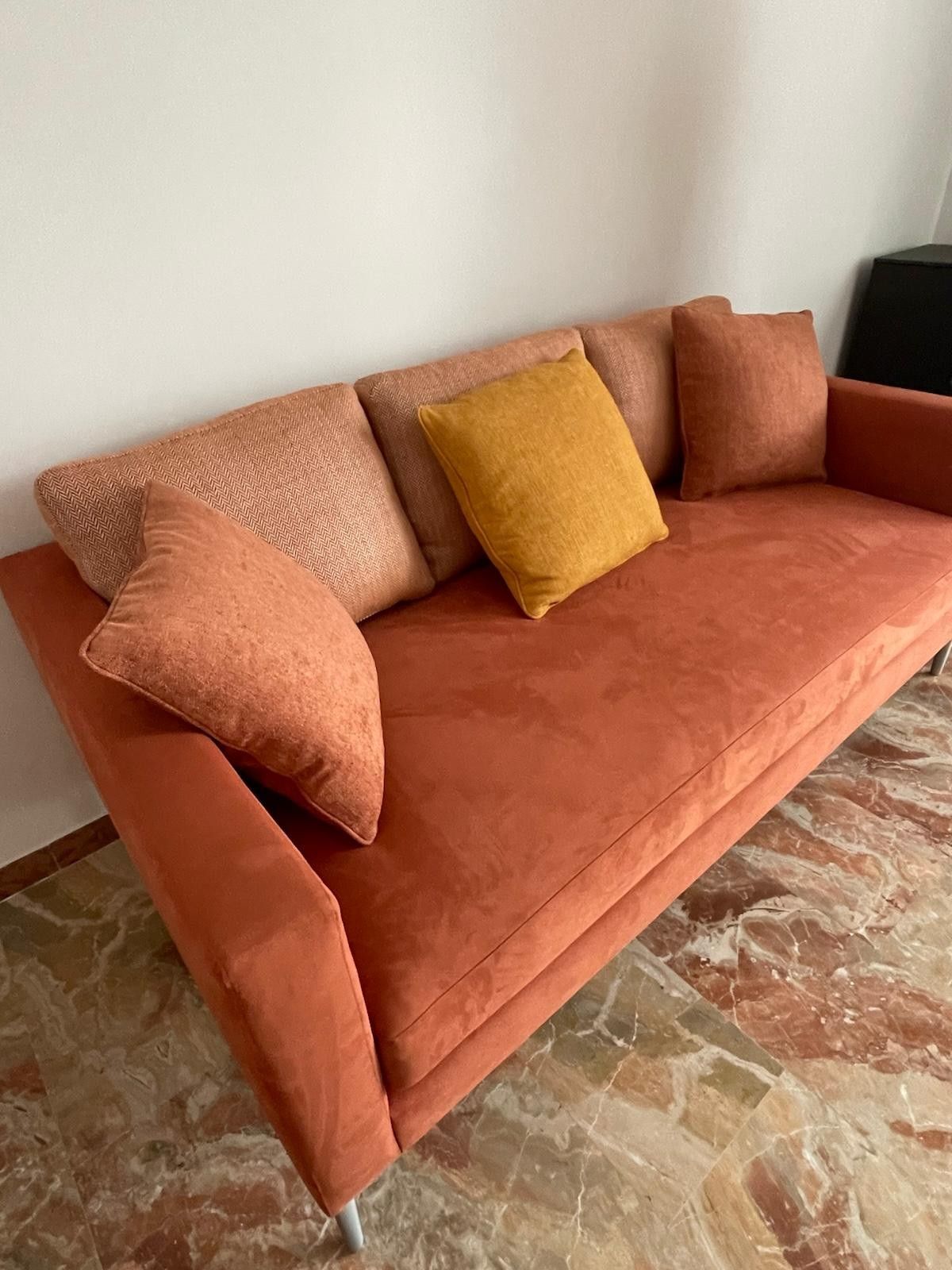 Divano verde con tre cuscini e cuscini decorativi a fantasia. Dettagli con bordino beige.