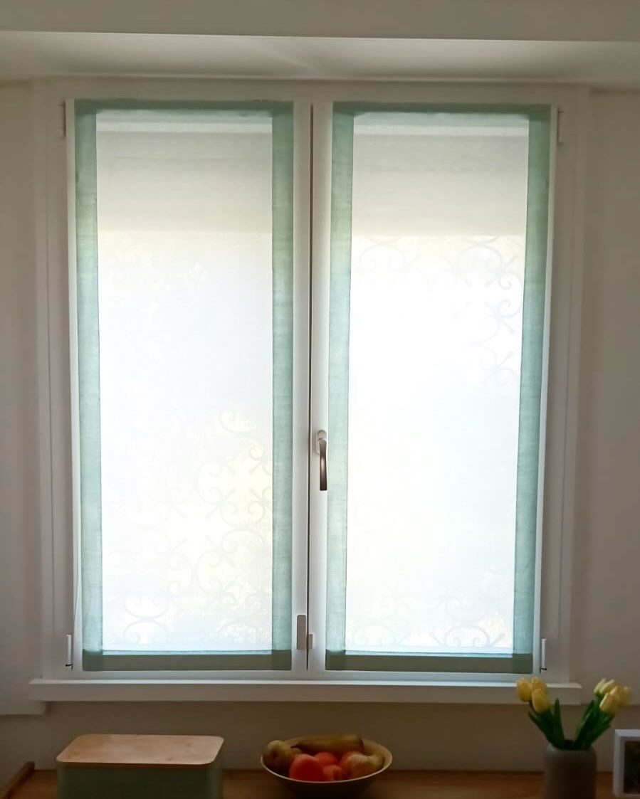 Finestra a due ante con tende bianche trasparenti, finiture verde chiaro e davanzale.