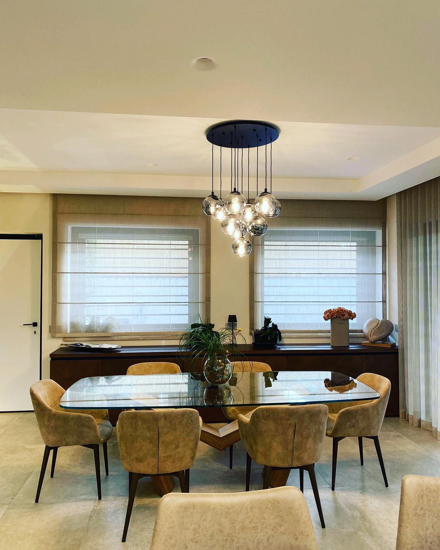 Sala da pranzo con tavolo in vetro, sedie imbottite, lampadario moderno e credenza in legno scuro.