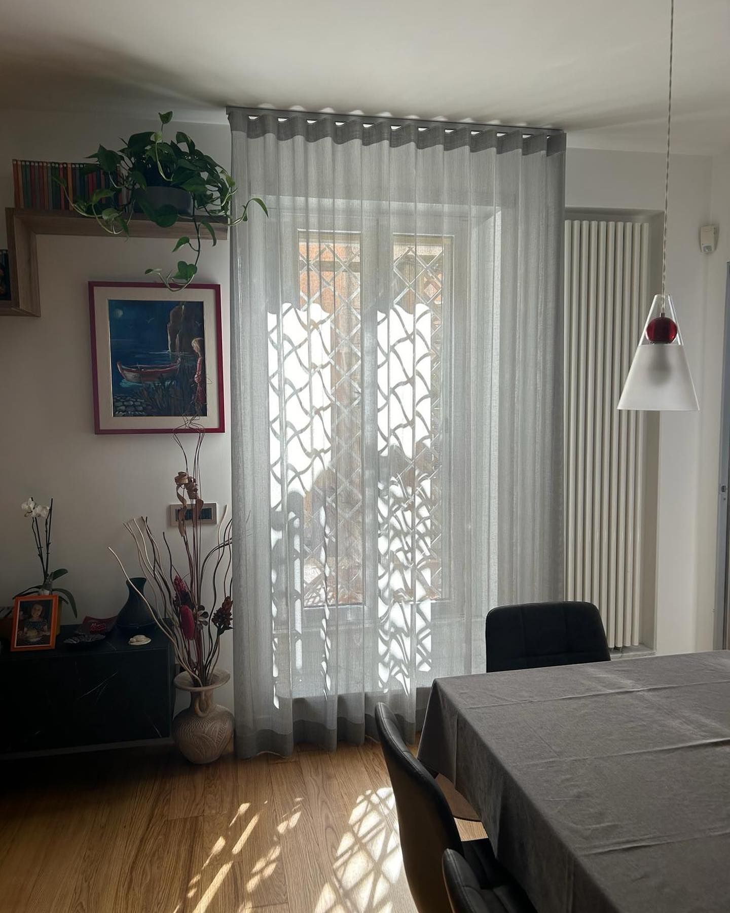Soggiorno illuminato dal sole con tende trasparenti, tavolo, piante e decorazioni murali.