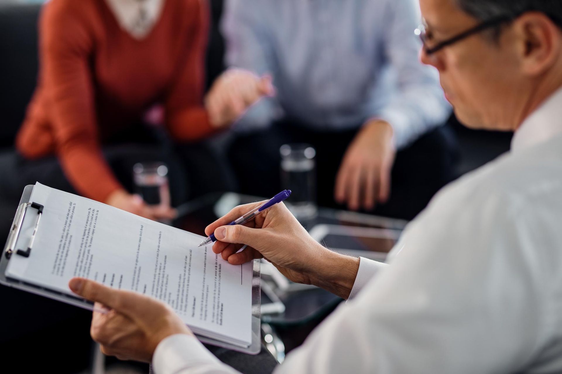 Person filling out a form on a clipboard during a meeting with seated colleagues