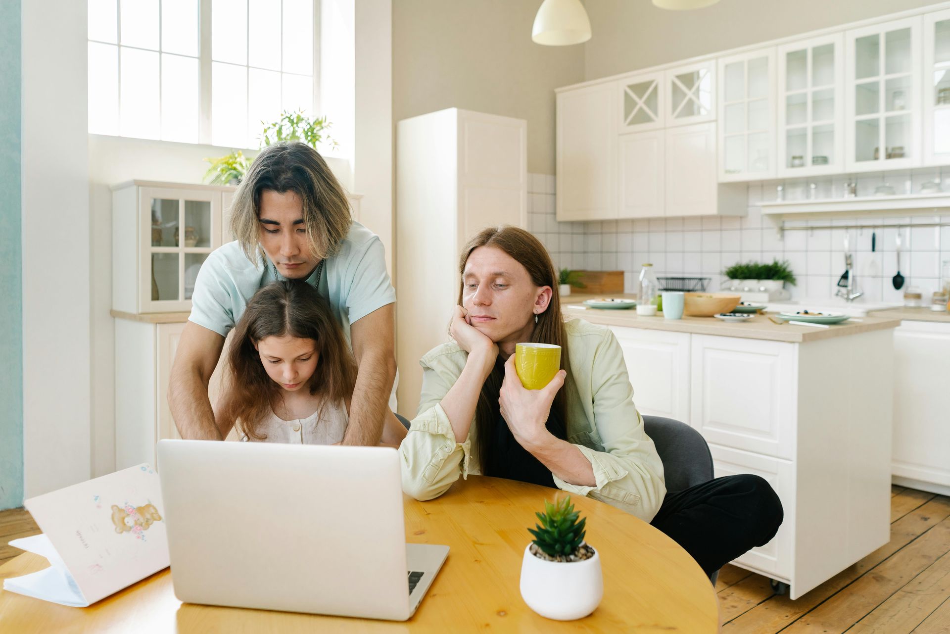 Three people gathered around a laptop at a kitchen table in a bright white kitchen.