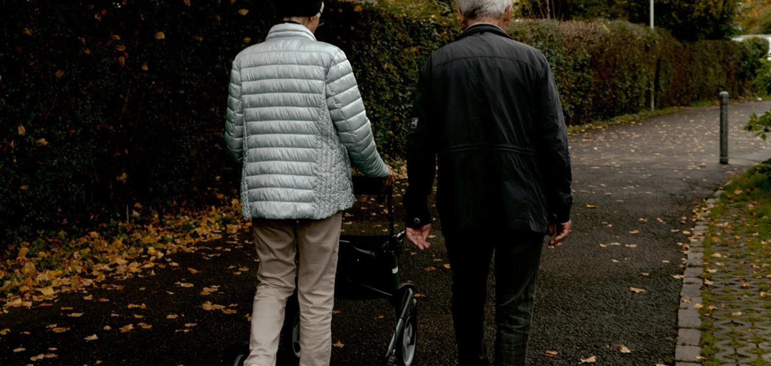 Older couple walking together on an autumn path, one using a rolling walker among fallen leaves.
