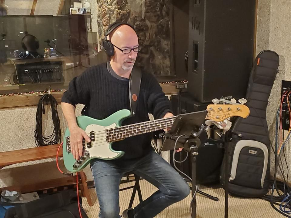A person wearing headphones plays a seafoam green bass guitar in a music studio, seated with a tablet on a stand.
