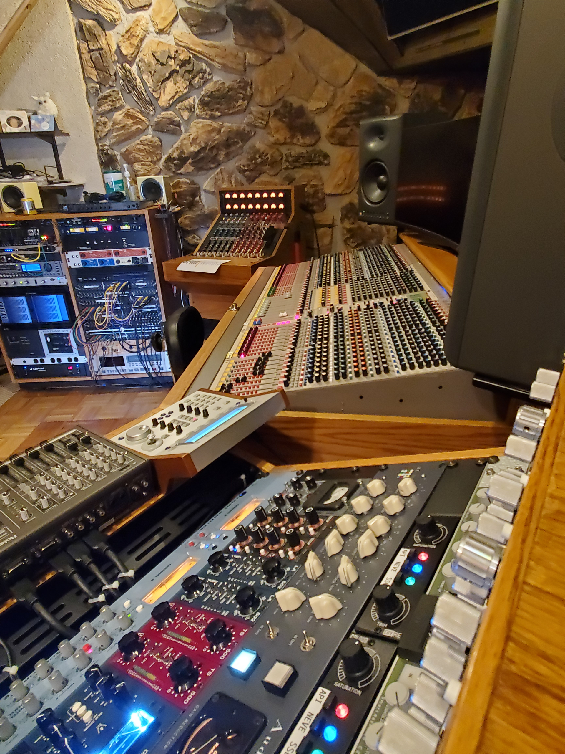 A recording studio control room featuring a large mixing console, outboard gear racks, and a stone wall background.