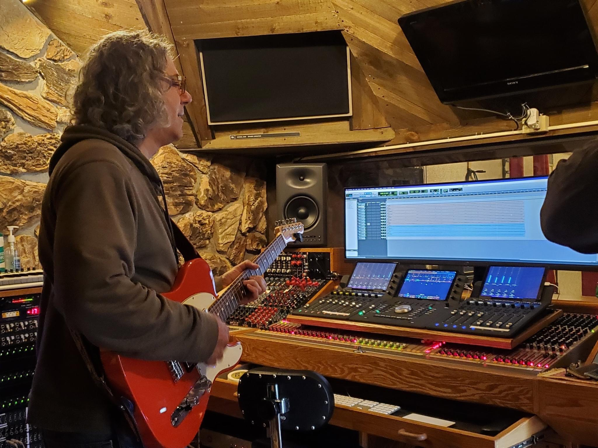 A person plays a red electric guitar in a studio, standing before a large mixing console with computer monitors.