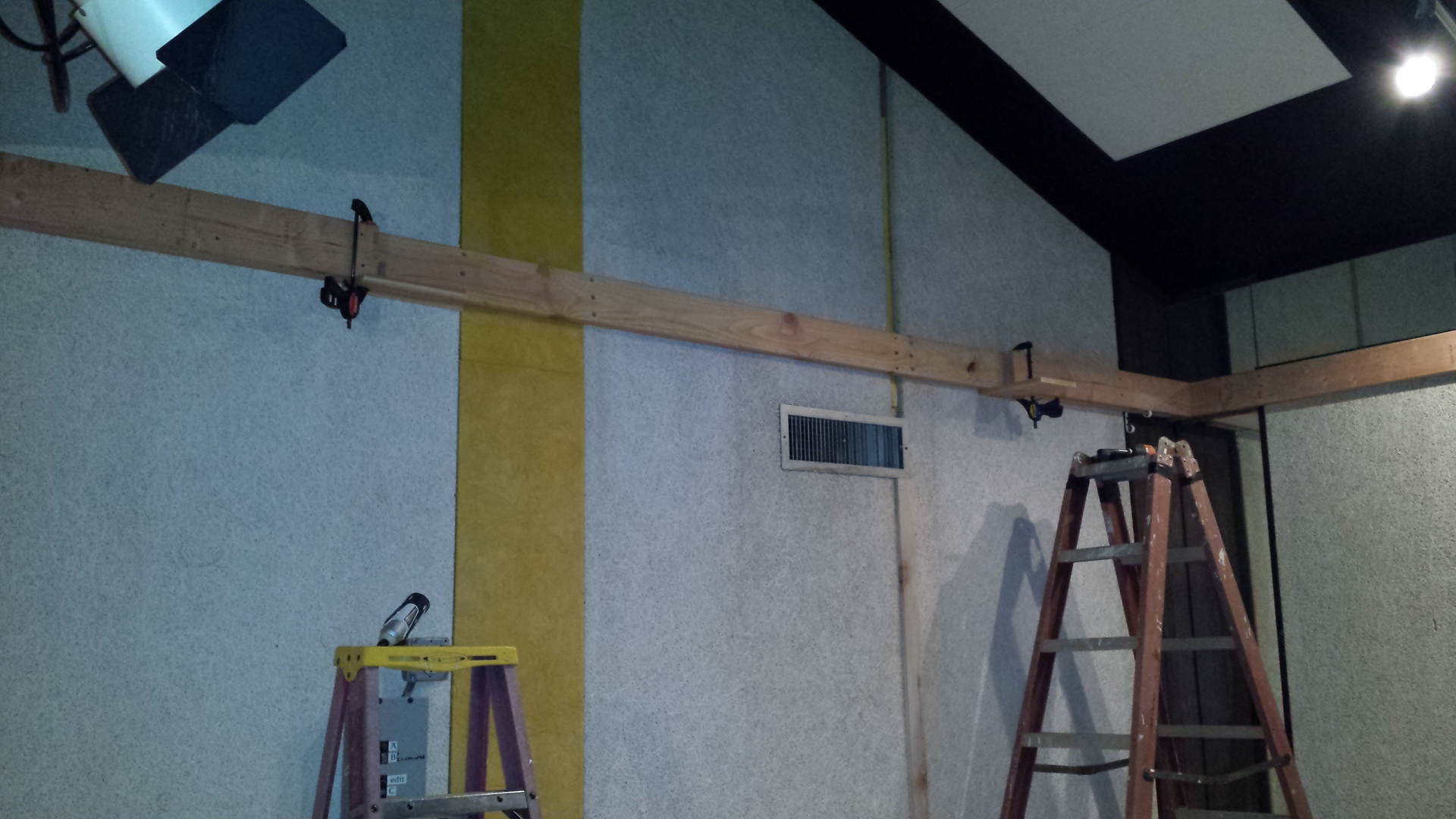 A studio space with a wooden support beam mounted across light-textured walls, featuring two ladders and overhead lights.