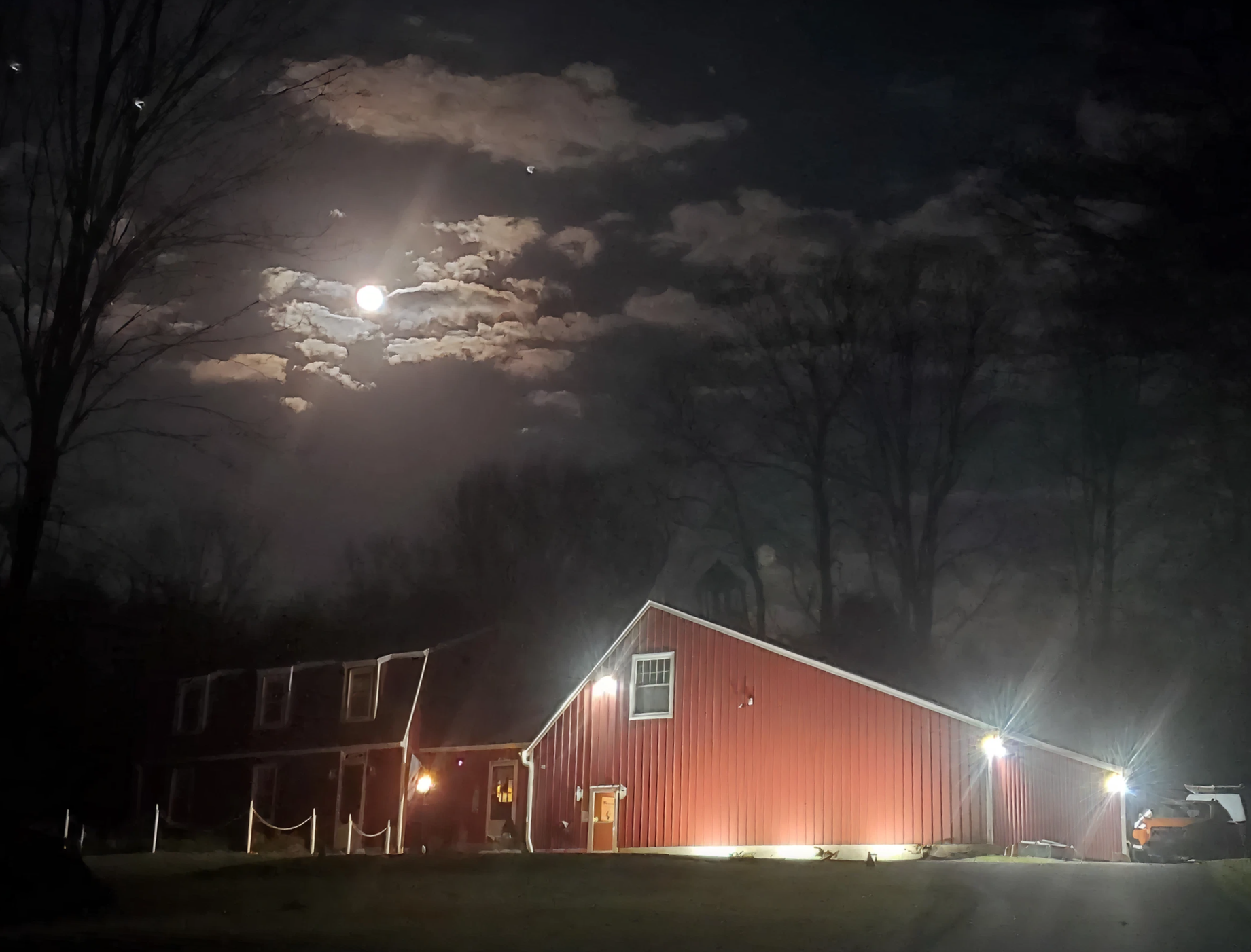 A red barn with lit exterior lights sits in a dark, rural landscape under a night sky with a bright moon and clouds.