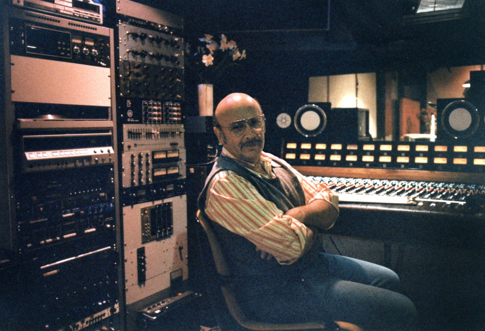 A person with a mustache and glasses sits with arms crossed in front of a recording studio mixing console and equipment.